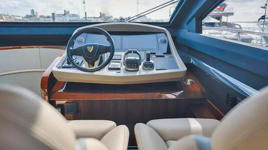 Inside a luxury boat, featuring a steering wheel, control panel with gauges, and plush leather seats. Sunny harbour view visible.